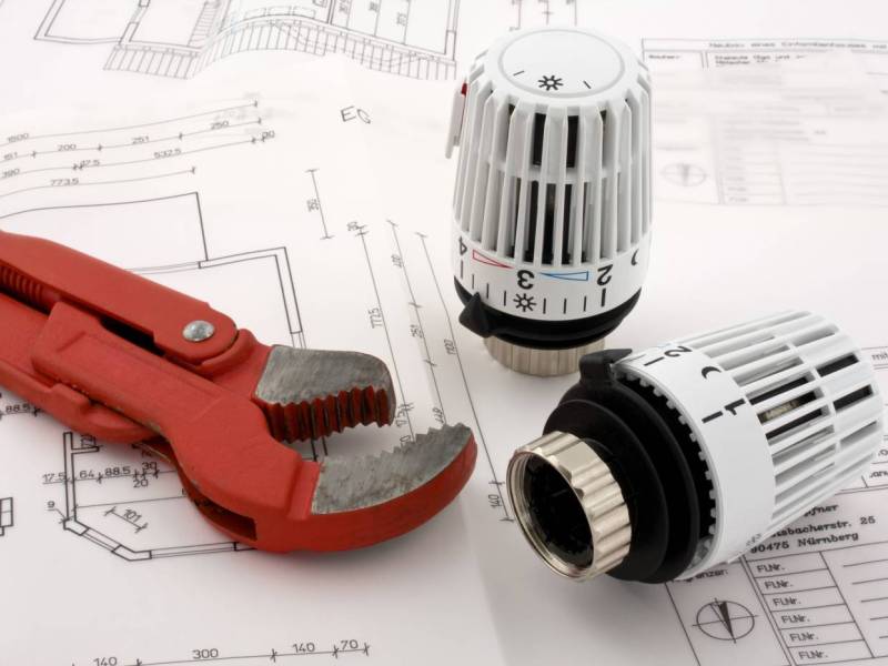 Ein rotes Rohrzangen-Werkzeug liegt auf einem Plan, daneben zwei Heizkörperthermostate.